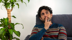 Handsome male model feels concerned puzzled lost in thoughts pondering making decision concept. Man wearing a winter sweater and thinking while sitting on couch home.